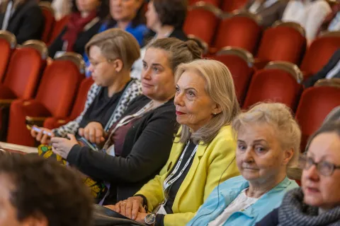 Na zdjęciu widać grupę osób siedzących w rzędach czerwonych foteli podczas konferencji. Uczestniczki, w różnym wieku, uważnie słuchają prelekcji – niektóre mają skupiony wyraz twarzy, inne subtelnie się uśmiechają. W centrum kadru wyróżnia się kobieta w żółtej marynarce, patrząca z zainteresowaniem w stronę sceny.