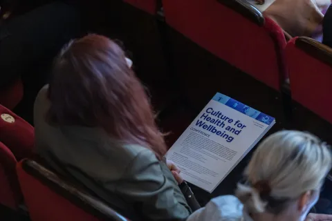 Na zdjęciu uczestniczka konferencji **"Culture for Health and Wellbeing"** siedzi w czerwonym fotelu audytoryjnym, trzymając w rękach program wydarzenia. Ujęcie wykonane z góry ukazuje jej skupienie oraz atmosferę konferencji, odbywającej się w eleganckiej sali.
