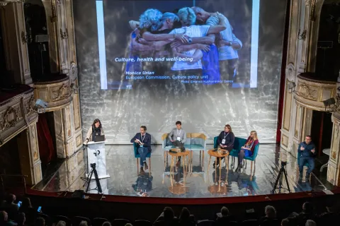 Zdjęcie przedstawia panel dyskusyjny na temat „Kultura, zdrowie i dobrostan” w eleganckim teatrze. Na scenie znajduje się prelegentka przy podium oraz czworo panelistów siedzących na krzesłach. Za nimi wyświetlona jest prezentacja z obrazem grupy ludzi przytulających się. W tle widać bogato zdobione wnętrze teatru, a na widowni znajdują się uczestnicy konferencji. Po prawej stronie sceny mężczyzna w czarnym ubraniu pokazuje gest aprobaty (kciuki w górę).