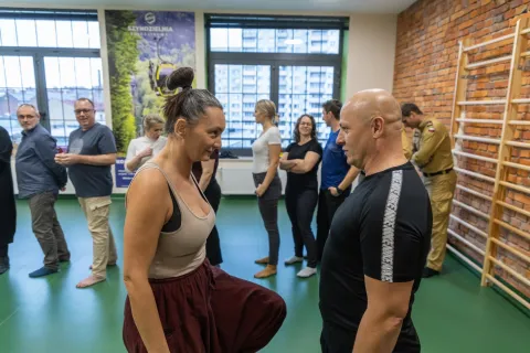 Na zdjęciu uczestnicy **warsztatów dla służb mundurowych** prowadzonych przez **Kasię Szewciów** biorą udział w ćwiczeniach opartych na ruchu i świadomości ciała. W centrum kadru kobieta i mężczyzna utrzymują intensywny kontakt wzrokowy, wykonując zadanie wymagające równowagi i skupienia. W tle inni uczestnicy ćwiczą w parach, w przestronnej sali z zieloną podłogą i ceglanymi ścianami, co tworzy atmosferę swobodnej, ale uważnej pracy.