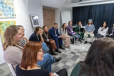 Na zdjęciu uczestnicy warsztatów z Heather O'Donnell siedzą w kręgu w kameralnym, artystycznym wnętrzu. Grupa składa się z osób w różnym wieku, które słuchają z zaangażowaniem i skupieniem. Na ścianach widoczne są obrazy i fotografie, a w tle znajduje się tablica z zapisanymi tematami. Atmosfera sprzyja otwartej dyskusji i twórczej wymianie myśli.