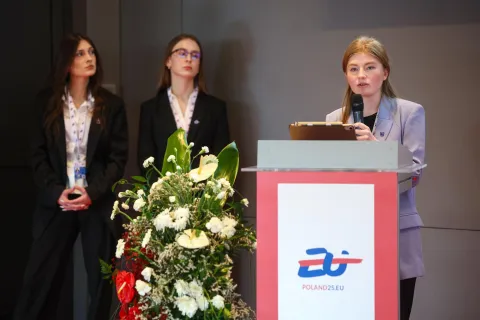 Młoda kobieta przemawia przy mównicy z logo Poland25.eu, trzyma mikrofon i tablet; obok niej stoją dwie uczestniczki wydarzenia w ciemnych garniturach. W tle kompozycja z białych i czerwonych kwiatów