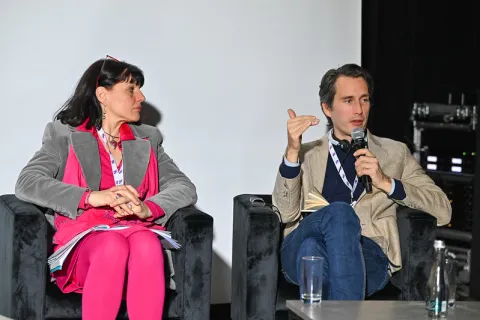 Na zdjęciu widzimy Nilsa Fietje i Monicę Urian siedzących obok siebie na czarnych fotelach podczas panelu dyskusyjnego. Nils Fietje, trzymając mikrofon i notatnik, gestykuluje, wyrażając swoją wypowiedź. Monica Urian słucha uważnie, z otwartym notatnikiem na kolanach. Oboje mają identyfikatory zawieszone na smyczach. W tle widać fragment sprzętu technicznego i neutralną białą ścianę. Atmosfera jest skupiona i profesjonalna.