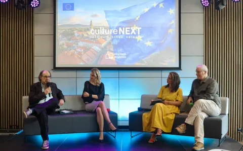 Na zdjęciu czterech ekspertów siedzi na scenie podczas panelu dyskusyjnego w nowoczesnej sali konferencyjnej. W tle znajduje się duży ekran wyświetlający grafikę z flagą Unii Europejskiej oraz napisem „culture NEXT – candidate cities network” i logiem Książnicy Beskidzkiej. Panelista po lewej stronie trzyma mikrofon i mówi, a pozostali uczestnicy słuchają go z zainteresowaniem. Oświetlenie sceniczne dodaje dynamiki wydarzeniu.