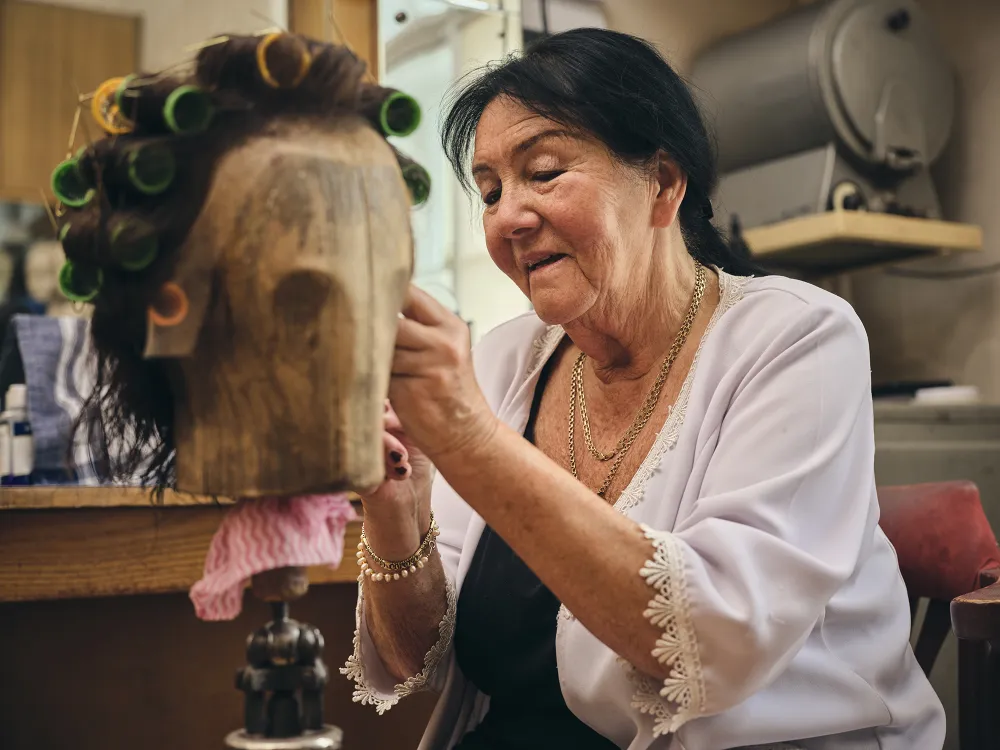 Starsza kobieta z uśmiechem pracuje nad peruką umieszczoną na drewnianej główce. Włosy mają założone wałki, a scena rozgrywa się w przytulnym, rzemieślniczym zakładzie.