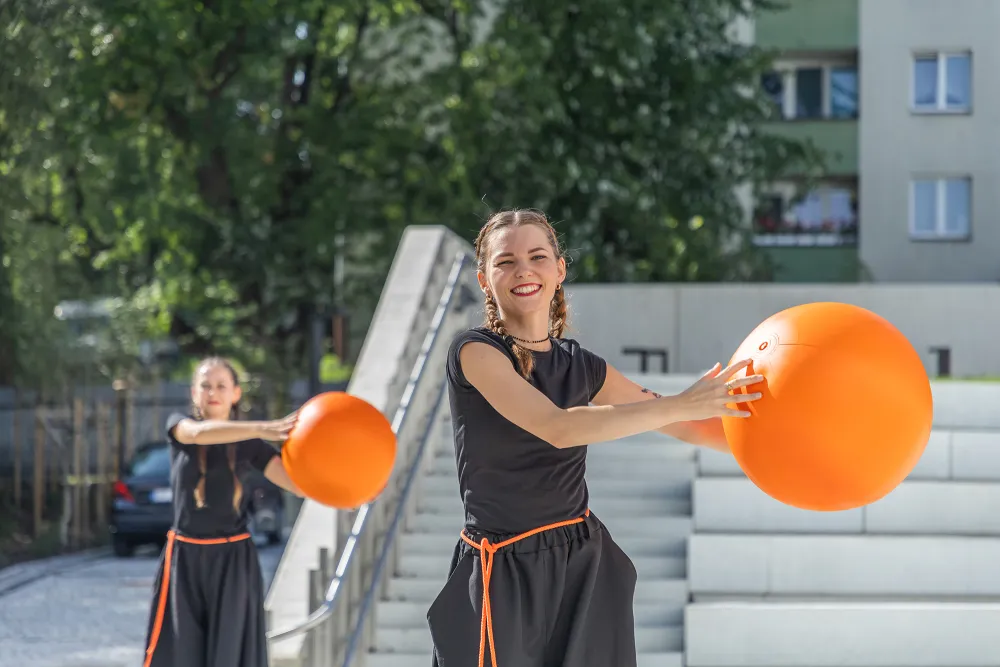 Dwie młode kobiety w czarnych strojach z pomarańczowymi akcentami tańczą na szczudłach na świeżym powietrzu, trzymając duże, pomarańczowe piłki. Na twarzy jednej z nich widać radosny uśmiech. W tle schody, drzewa i fragment budynku mieszkalnego.